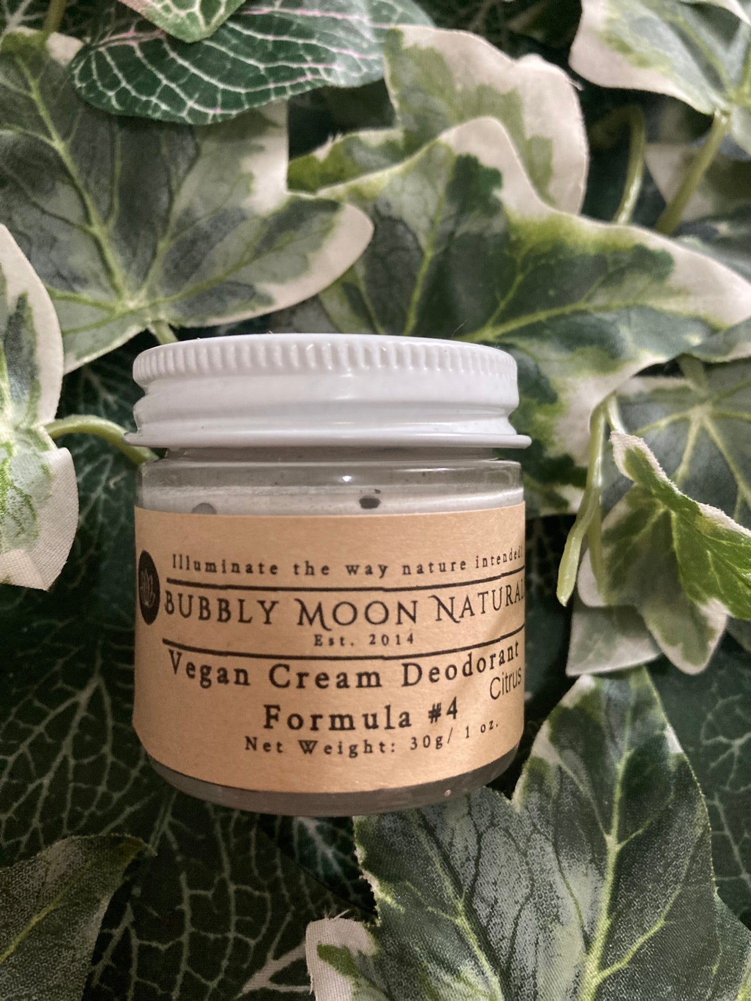 Vegan Cream Deodorant tan label with white metal top in glass jar laying on artificial leaf grass. Bubbly Moon Naturals name and product info is displayed on label.