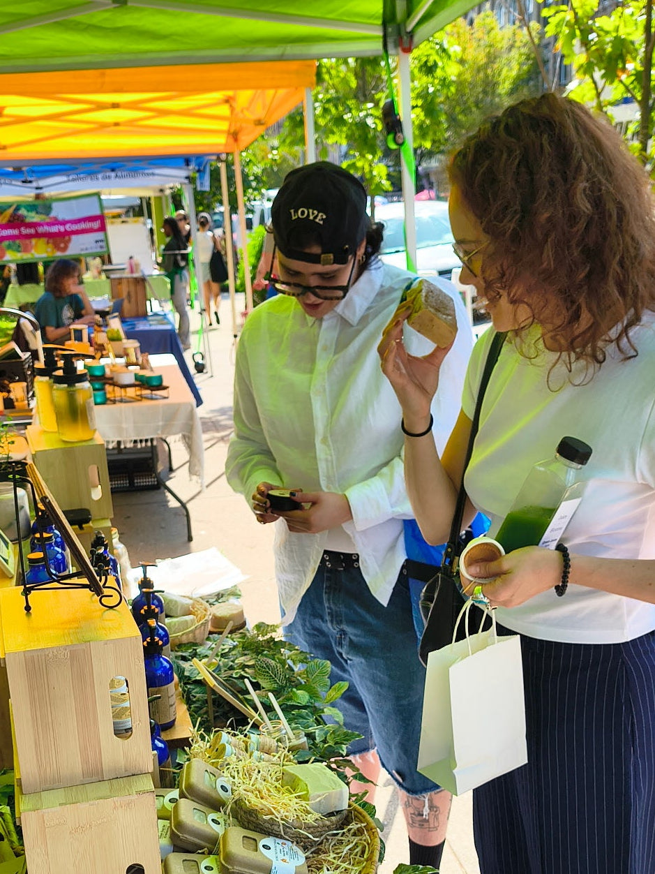 Photo of customers smelling soap and checking out at the Bubbly Moon Naturals' Vendor Stall at the Bushwick Farmer's Market. Two people in white shirts and blue jeans and striped pants. The one on the left is viewing the Salve no. 1 and the other is smelling Artisan Bar No.  under a green tent with green foliage and wood boxes on the table. 