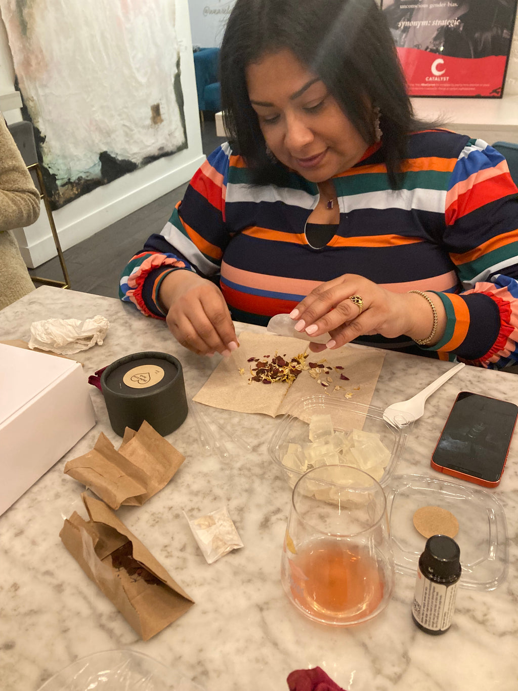 Participant pouring herbal mixture together before adding to her melt and pour soap and scrub. Measuring spoons, herbs and boxes are on the table. 