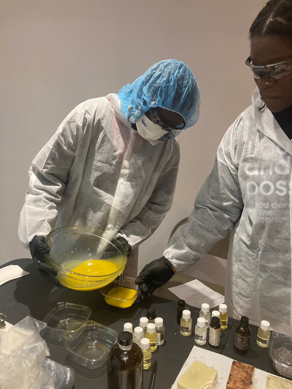 Cold-process soap making experience with participants pouring soap into molds wearing protective personal equipment with scents and materials on the table.